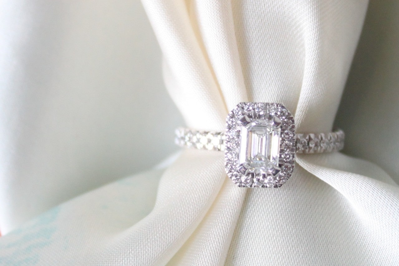 an emerald cut, halo set, white gold engagement ring with white fabric threaded through it