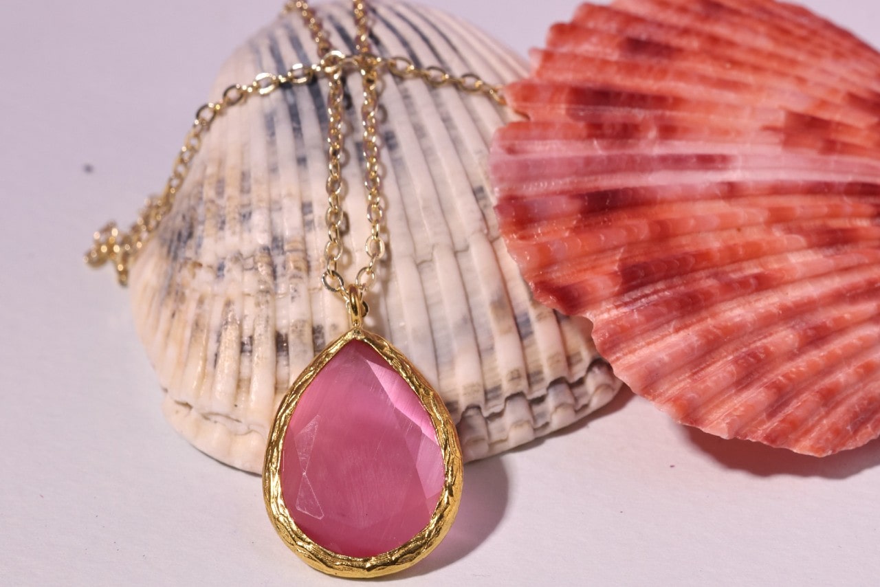 a yellow gold pendant necklace featuring a large pink pear shaped gemstone lying on seashells