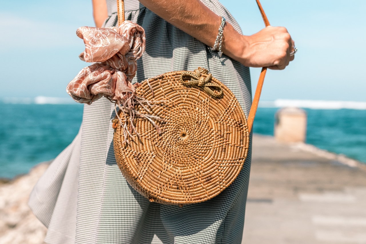 a woman holding a wicker purse on the beach and wearing a silver chain bracelet