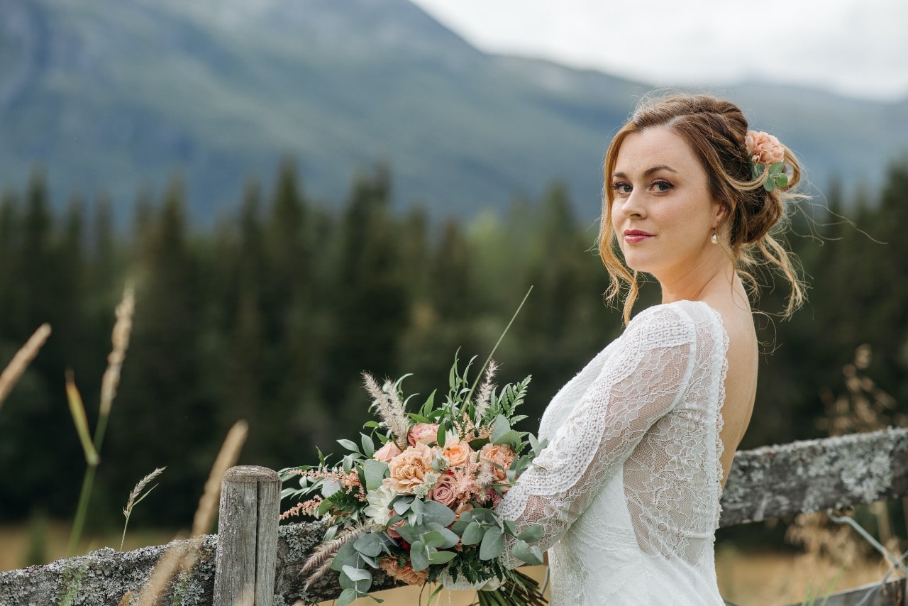 A bride leans against a rustic wooden fence in a field with mountains.