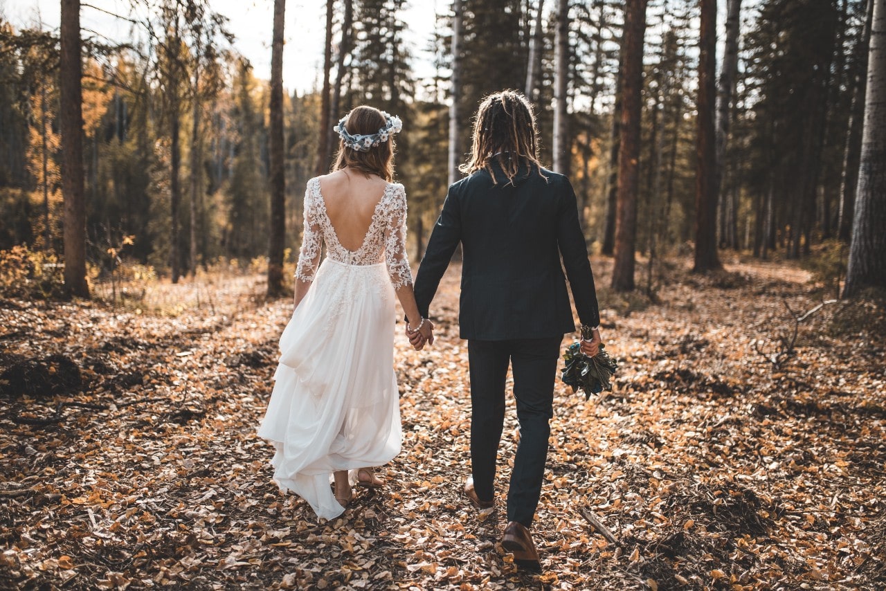 A groom and bride walk through a fall forest while holding hands.