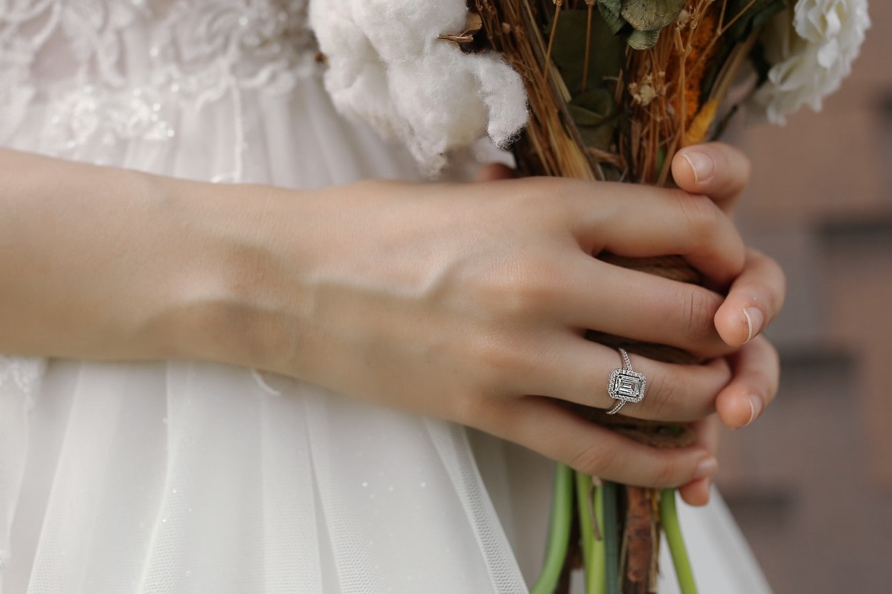 a bride holding a bouquet of flowers and wearing an emerald cut halo engagement ring