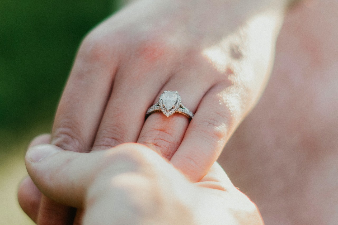 two hands holding, one of which is wearing a pear shape halo engagement ring