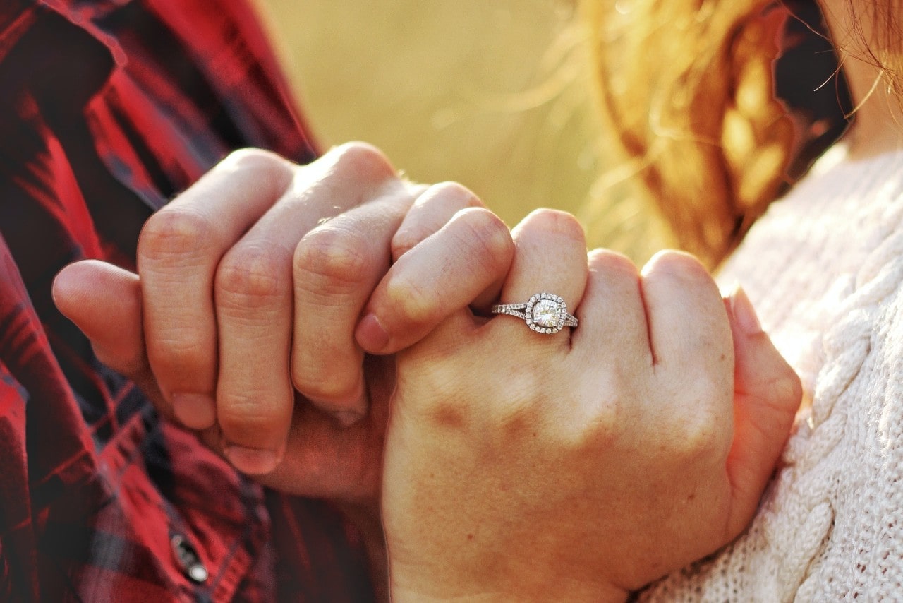 a couple intertwining pinkies, the woman wearing a cushion cut, halo engagement ring