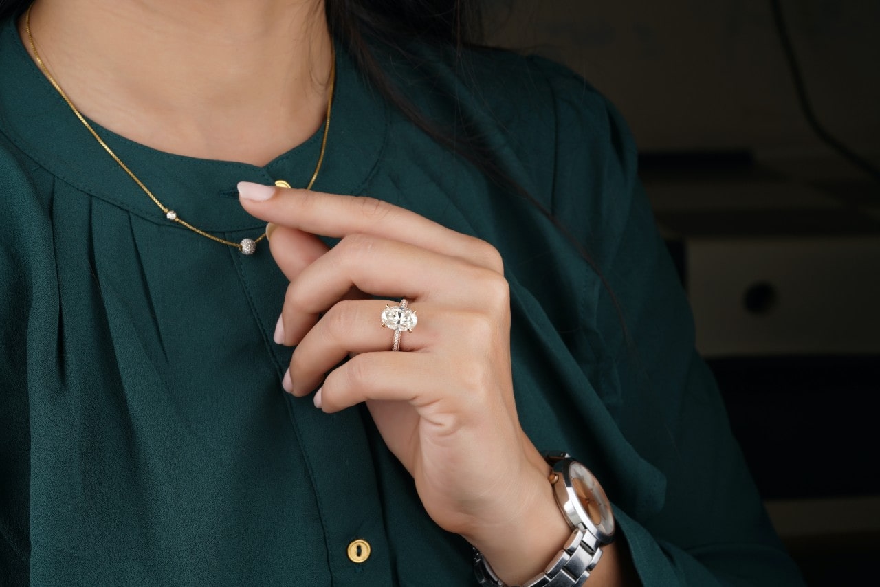 A side stone oval cut engagement ring on a woman’s hand as she touches her necklace.