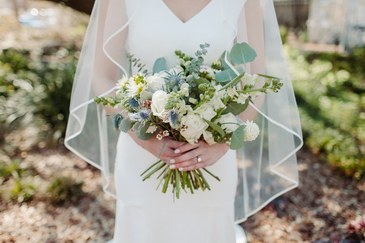 A bride holding a bouquet wears her engagement ring while standing outside.