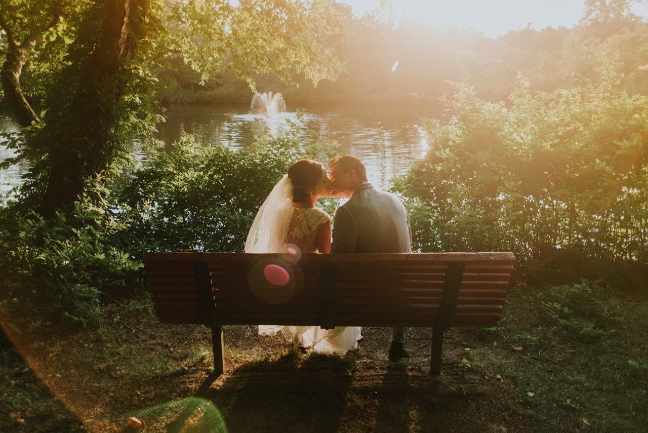 A newly-wed couple sits on a bench by the lake at a park and celebrate with a kiss.